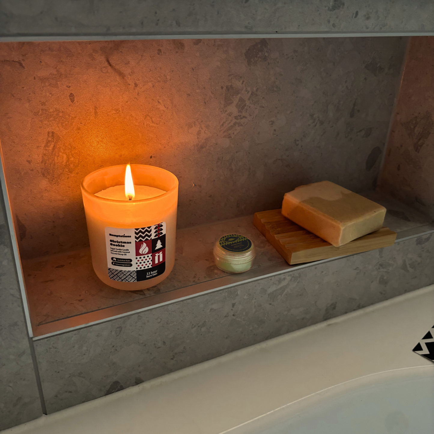 Candle with a label on a shelf next to soap and a jar, with a warm glow.