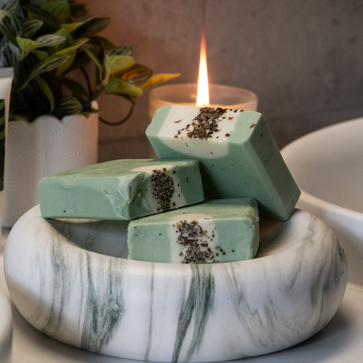 Green soap bars in a marble bowl with candles and bottles on a bathroom counter.